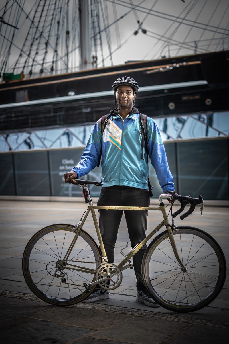 A cyclist in helmet and blue jacket standing with a road bike, tall ship rigging behind